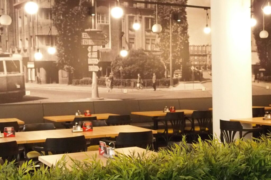 Moderní interiér restaurace s dřevěnými stoly, černými židlemi, závěsnými svítidly a zelenými rostlinami v popředí. Zadní stěnu pokrývá velká černobílá nástěnná malba s motivem městské krajiny. Zažijte příjemnou atmosféru restaurace Gurmán Dukelská – ideální místo pro skvělý oběd v Hradci Králové. Na snímku nejsou žádné osoby.
