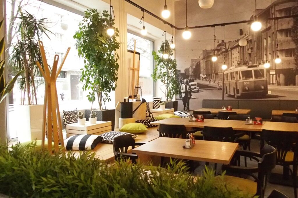 Moderní kavárna s dřevěnými stoly, černými židlemi, zelenými rostlinami a velkou nástěnnou malbou zachycující retro pouliční scénu. Prostorem proniká přirozené světlo velkými okny a nad hlavami visí kulatá stropní svítidla. Útulné polštáře zdobí posezení a vytvářejí ideální místo, kde si můžete vychutnat svůj oběd Prima nebo si odpočinout při čekání na rozvoz obědů v Hradci Králové od Gurmán Dukelská.