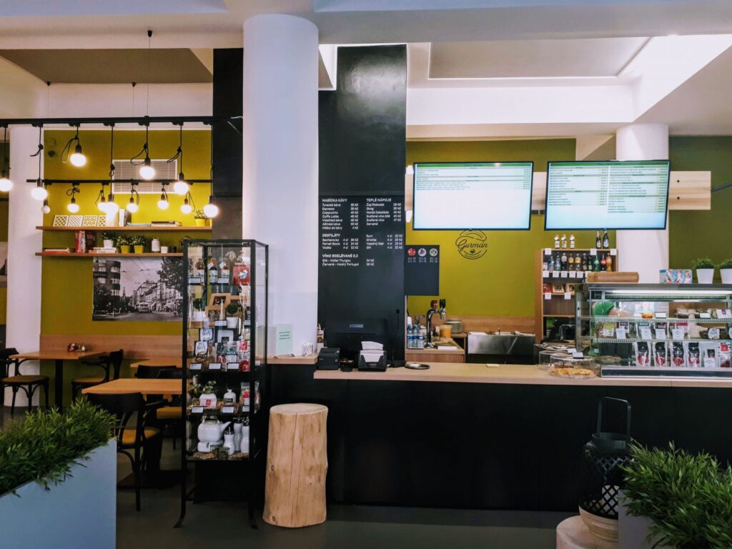 Moderní interiér kavárny s černým pultem, digitálními tabulemi s denním menu v Hradci Králové, výstavními policemi s produkty, dřevěnými stoly a židlemi, závěsnými svítidly a zelenými akcentními stěnami s rostlinami v popředí – ideální místo pro vychutnání „Prima oběda“ v restauraci Gurmán Dukelská.