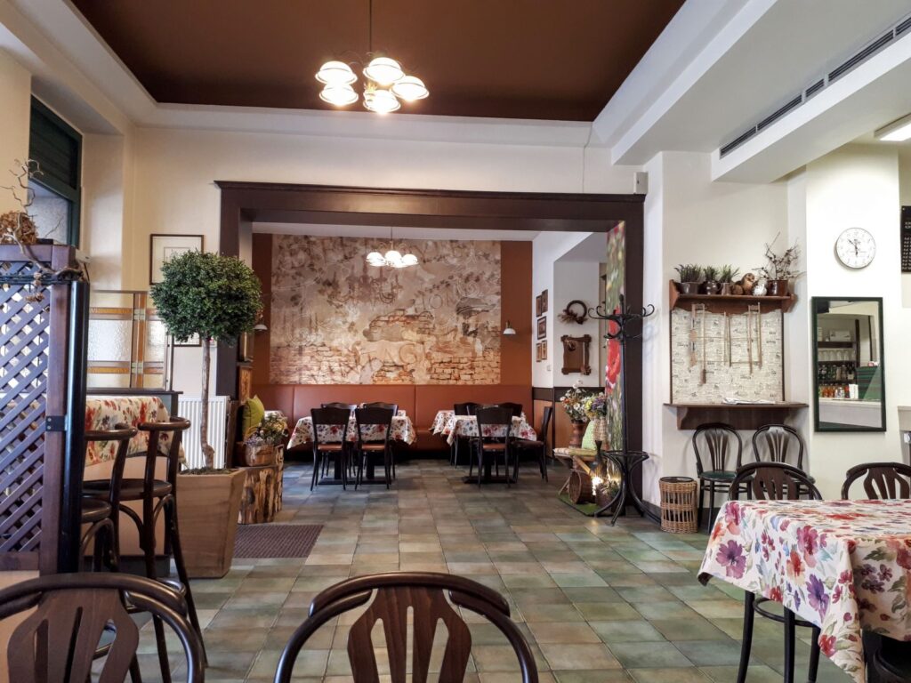 Útulný interiér restaurace s prázdnými stoly pokrytými květinovými ubrusy, dřevěnými židlemi, nástěnnou malbou na zadní stěně, dekorativními rostlinami a příjemným osvětlením. Prostředí je příjemné a rustikální – ideální pro vychutnání denního menu v Hradci Králové v restauraci Gurmán Gočárova nebo pro ty, kteří využívají rozvoz obědů v Hradci Králové.