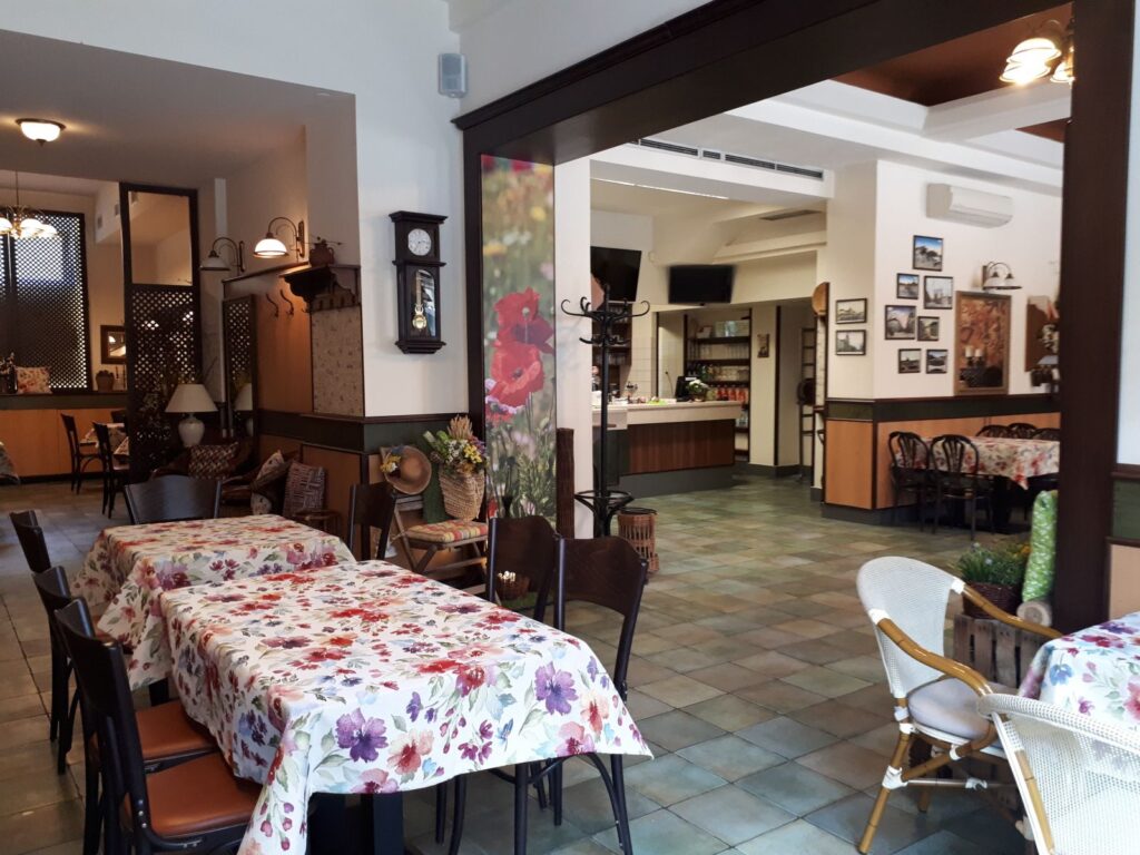 Útulná, prázdná restaurace s květinovými ubrusy, dřevěnými židlemi a vintage výzdobou. Stěny zdobí nástěnné hodiny a zarámované fotografie. Prostor je dobře osvětlený přirozeným i umělým světlem – je to příjemné prostředí, kde si můžete vychutnat oběd v Hradci Králové nebo si prohlédnout denní menu v Hradci Králové.