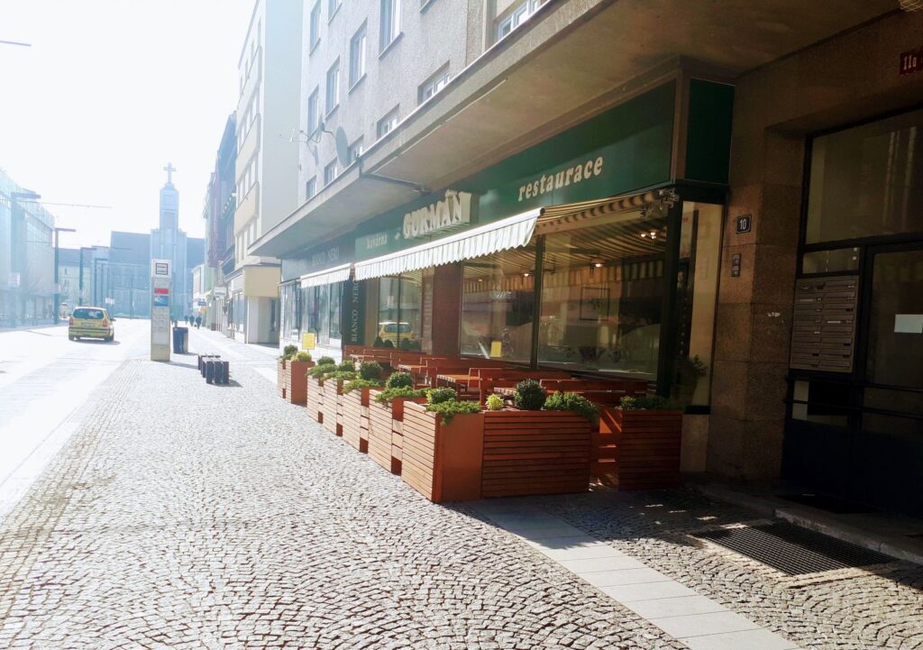 Klidná, sluncem zalitá městská ulice s restaurací, která nabízí venkovní dřevěné sezení a květináče; po obou stranách se tyčí budovy a v pozadí je vidět několik vozidel – ideální místo, kde si můžete vychutnat denní menu v Hradci Králové v restauraci Gurmán Dukelská nebo si dopřát „Prima oběd“ v okouzlujícím prostředí.