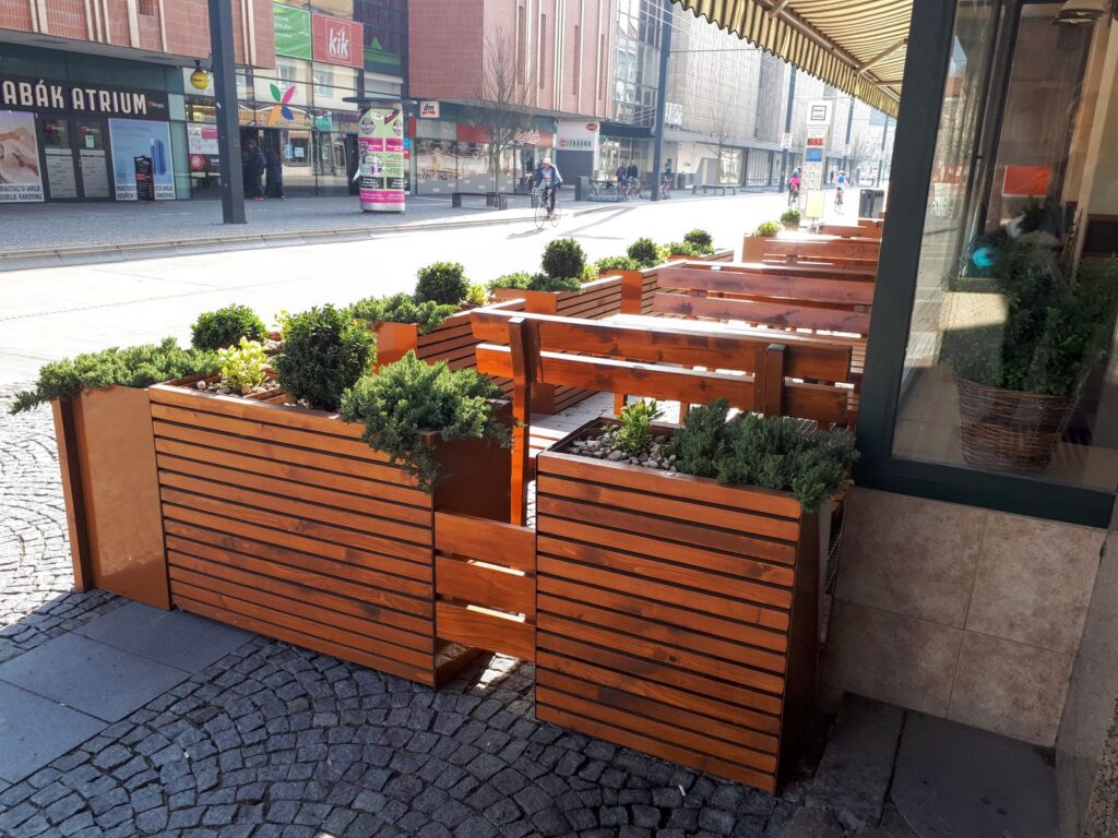 Venkovní posezení restaurace s dřevěnými stoly, lavicemi a květinovými truhlíky plnými zelených keřů, umístěné podél chodníku na městské ulici. V pozadí jsou vidět budovy, obchody a několik chodců. Tento příjemný prostor je ideální pro vychutnání obědů v Hradci Králové z oblíbeného denního menu restaurace Gurmán Dukelská a zároveň pro vnímání živé městské atmosféry.