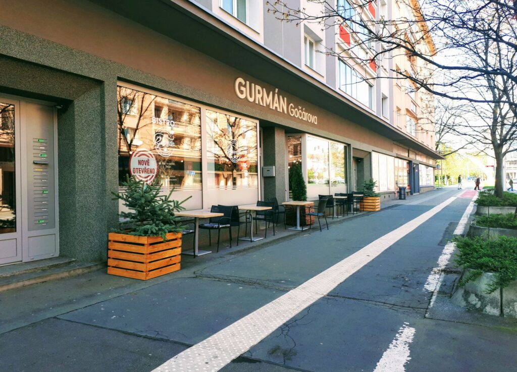 Venkovní posezení restaurace Gurmán Gaštanova se stoly, židlemi a malými borovicemi v dřevěných květináčích podél chodníku pod řadou oken – ideální místo, kde si můžete vychutnat „Prima oběd“ nebo ochutnat některé z nejlepších obědů, které Hradec Králové nabízí.