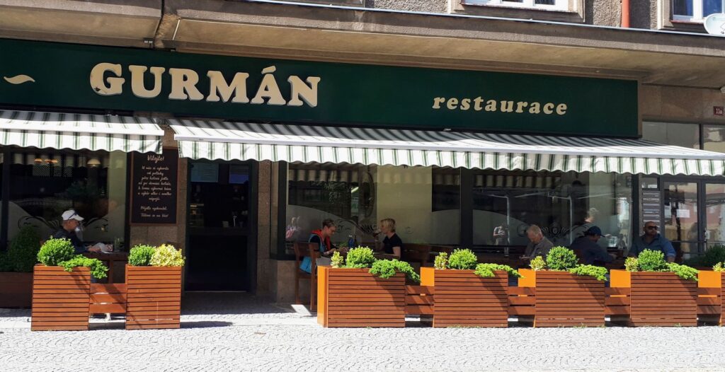 Venkovní posezení v restauraci Gurmán – známé také jako Gurmán Gočárova – nabízí hostům možnost stolovat pod pruhovanou markýzou, přičemž slunnou terasu lemují dřevěné květináče s zelenými keři. Vychutnejte si živou atmosféru na místě, nebo využijte jejich službu rozvozu obědů v Hradci Králové a vychutnejte si „Prima oběd“ v pohodlí svého domova.