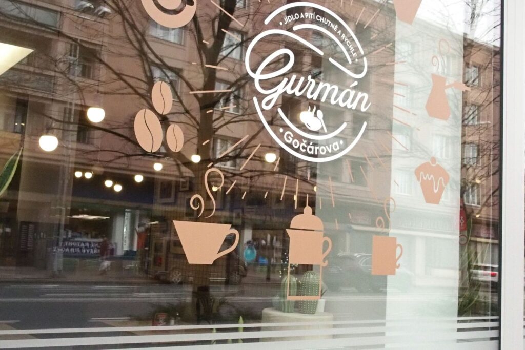 V okně kavárny s nálepkami s motivy kávy – včetně šálků, zrn a francouzského lisu – se odráží ulice, stromy a budovy venku. Logo kavárny „Gurmán Gočárova“ je výrazně vystaveno vedle nenápadného odkazu na „Gurmán Dukelská“ a denního menu „Hradec Králové“ – což zdůrazňuje, že se jedná o ideální místo pro „Prima oběd“ v samém srdci města.
